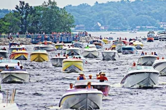 Numerous boats on a crowded lake with trees and spectators in the background.